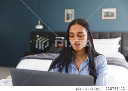 Young adult woman working on laptop in bedroom workspace with teal wall, potted plant and mug 128741412
