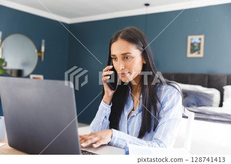 Woman talking on smartphone and typing on laptop at bedroom home office, with coffee mug 128741413