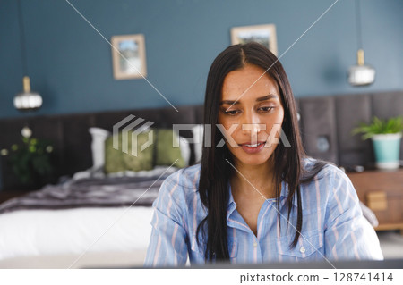 Young woman working on laptop at bedroom desk, surrounded by bed, plants and wall art 128741414
