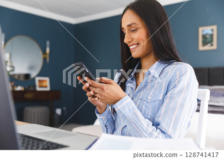 Young woman sitting at desk in modern home office using laptop and checking smartphone, copy space 128741417