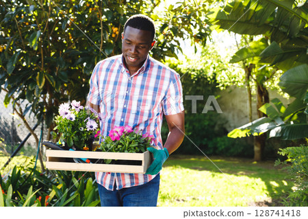African American man carrying crate of potted flowers and trowel in garden, wearing green gloves 128741418