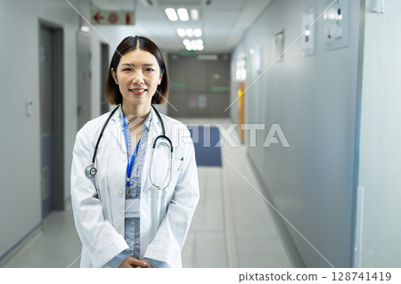 Asian woman standing and smiling in hospital hallway, wearing lab coat, stethoscope and ID badge Asian woman standing and smiling in hospital hallway, wearing lab coat, stethoscope and ID badge 128741419