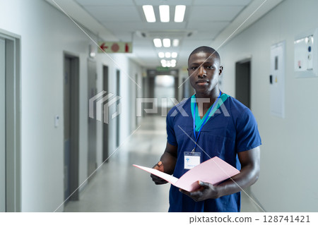 African American male nurse reviewing pink folder in hospital corridor wearing ID badge, copy space 128741421