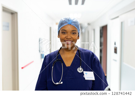 Female nurse smiling and standing in hospital corridor, wearing surgical cap, stethoscope, ID badge 128741442