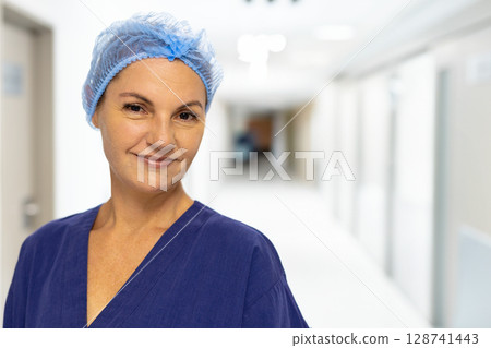 Female nurse standing and smiling in hospital corridor, wearing navy-blue scrub top and hairnet 128741443