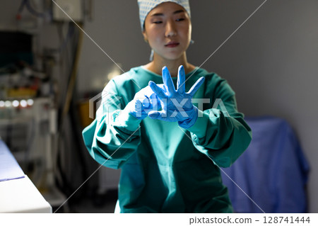 Young Korean woman surgeon pulling on blue gloves in dim operating room, with surgical drape 128741444
