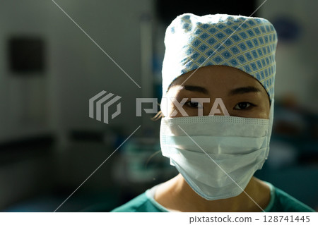 Asian female surgeon gazing at camera in hospital operating room, highlighting scrub cap and mask 128741445