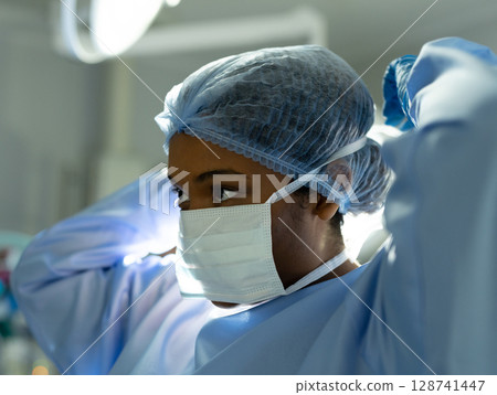Female surgeon adjusting mask straps and surgical cap in operating room, with bright overhead light Female surgeon adjusting mask straps and surgical cap in operating room, with bright overhead light 128741447