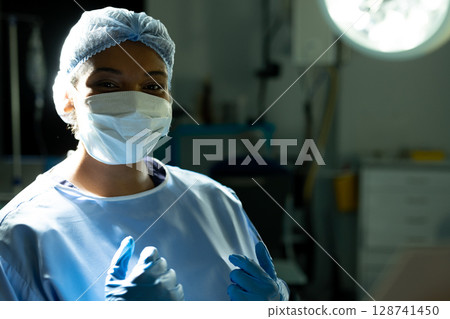 Diverse female surgeon adjusting latex gloves in hospital operating room, with gown cap mask 128741450