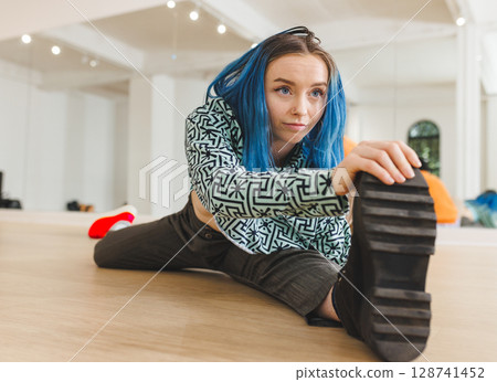 Non-binary person stretching leg wide on dance studio floor, reaching boot sole near studio mirror 128741452