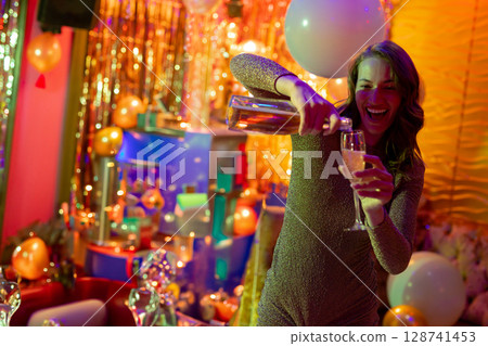 Woman pouring champagne into flute in party room, with balloons and gift boxes, copy space Woman pouring champagne into flute in party room, with balloons and gift boxes, copy space 128741453