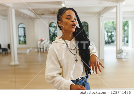 African American woman demonstrating dance moves in bright studio, with mirror wall, hoop earrings 128741456