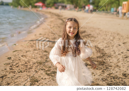 Cute beautiful long-haired girl in a white dress runs along the beach Cute beautiful long-haired girl in a white dress runs along the beach 128742082