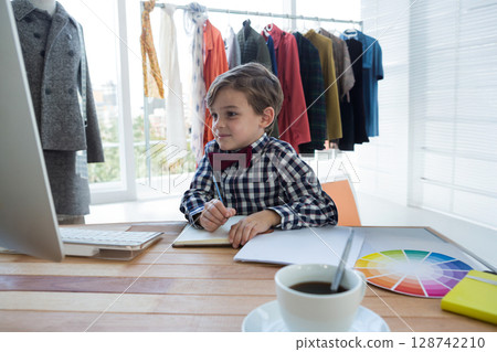 Boy child sketching in notebook in home studio at wooden desk, with computer monitor and keyboard 128742210