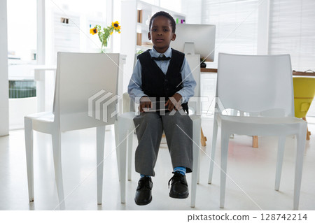 young African American boy sitting on plastic chair in study room, holding tablet near sunflowers 128742214