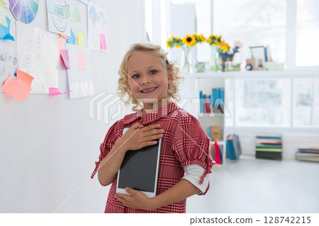 6-year-old girl standing next to sticky note covered wall in bright classroom space, holding tablet 128742215