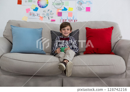 Child boy sitting on light grey couch in creative lounge, holding green mug with design sketches 128742216
