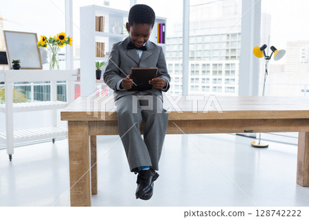 African American boy child sitting on table in modern studio, holding tablet near sunflowers 128742222