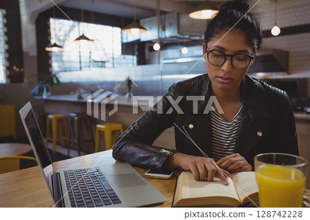 Indian woman reading open book at cafe table, tracing text with finger near laptop, copy space Indian woman reading open book at cafe table, tracing text with finger near laptop, copy space 128742228