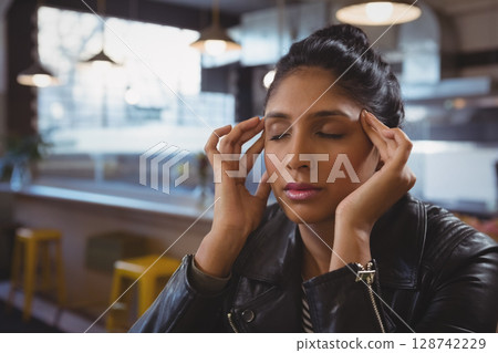 Indian woman sitting at table in modern cafe, pressing temples under industrial pendant lights Indian woman sitting at table in modern cafe, pressing temples under industrial pendant lights 128742229