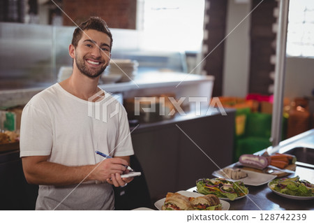 Young adult server taking order behind metal counter in deli, with pen and notepad, copy space 128742239