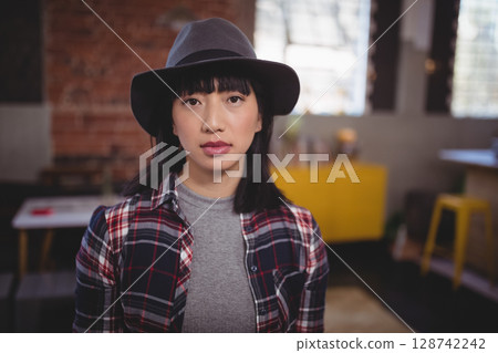 Mid-adult Asian woman standing in industrial style studio, wearing grey fedora, red plaid shirt 128742242