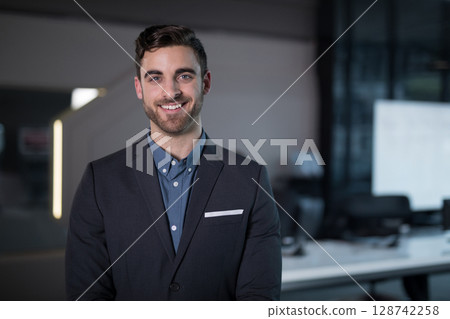Young adult man standing and smiling in open-plan office, displaying suit jacket and monitors 128742258