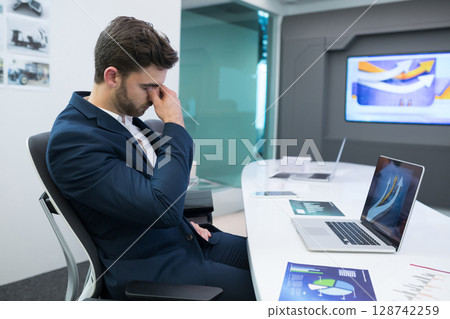 Young adult man pinching bridge of nose in conference room, with laptop, printed charts, copy space 128742259