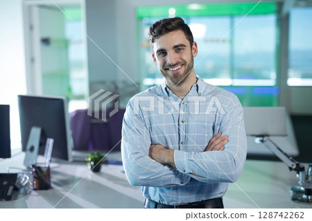 Man standing with arms crossed by desk in open plan office, surrounded by computer monitors, plant 128742262
