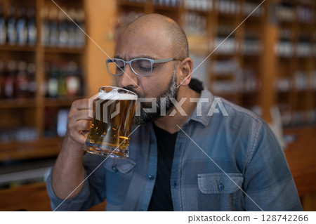 Indian man sipping beer from large glass mug at wooden bar counter, surrounded by bottled beers Indian man sipping beer from large glass mug at wooden bar counter, surrounded by bottled beers 128742266