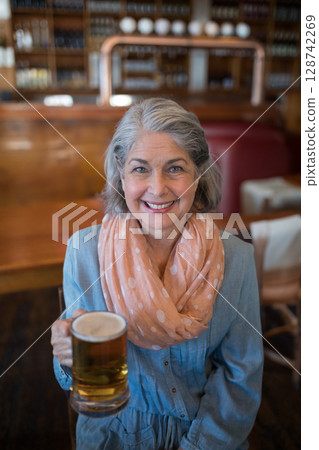 Senior woman sitting at wooden bar counter in cozy pub, holding frothy beer mug and smiling Senior woman sitting at wooden bar counter in cozy pub, holding frothy beer mug and smiling 128742269