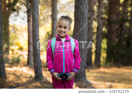 School-aged girl standing in pine forest wearing pink tracksuit, holding binoculars and backpack School-aged girl standing in pine forest wearing pink tracksuit, holding binoculars and backpack 128742270