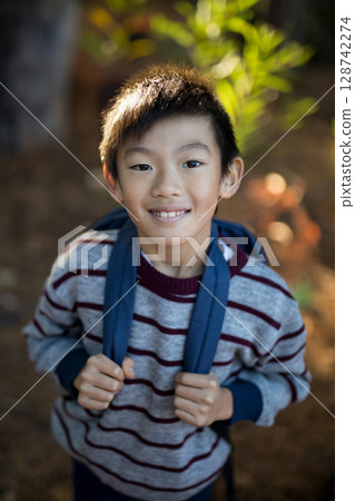 Asian boy smiling and standing on dirt path in wooded park, holding blue-strap backpack straps 128742274