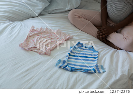 African American woman arranging infant onesies in bedroom on bed with sheets, pillows, copy space 128742276