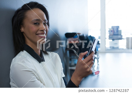 Young adult woman leaning against wall in lounge, using smartphone with suitcase behind, copy space 128742287