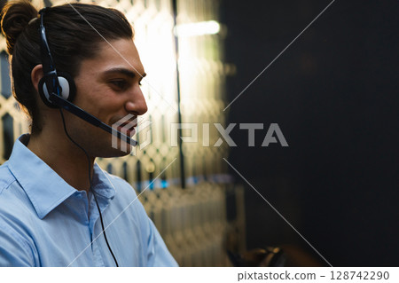 Young man speaking through over-ear headset in call center, with honeycomb partition, copy space Young man speaking through over-ear headset in call center, with honeycomb partition, copy space 128742290