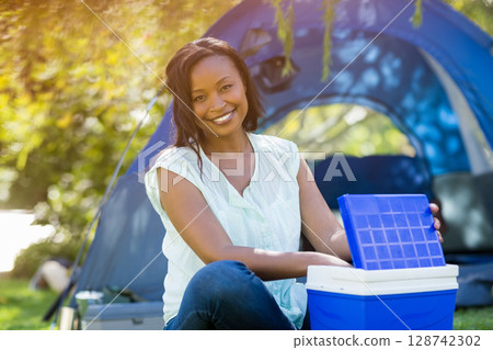 African American woman sitting on grass at campsite near blue dome tent, lifting cooler lid 128742302