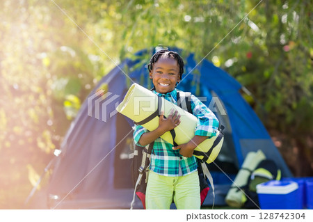 African American woman standing in forest campsite holding mat, wearing backpack with cooler box 128742304