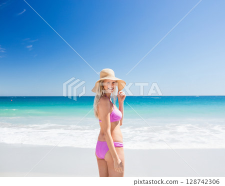 Young woman standing in profile on sandy beach, with pink bikini and wide?brimmed straw sun hat 128742306