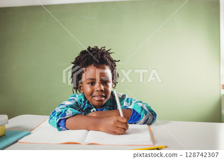 African American boy writing with pencil in notebook on white desk at school with green chalkboard 128742307