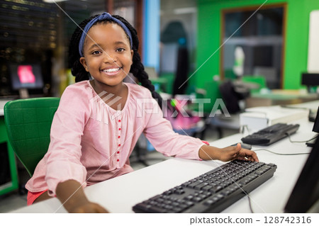 Teenage African American girl sitting at white desk in modern lab, reaching toward black keyboard 128742316