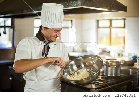 Young male chef preparing dish in restaurant kitchen, using stainless steel bowl and gas stove 128742331