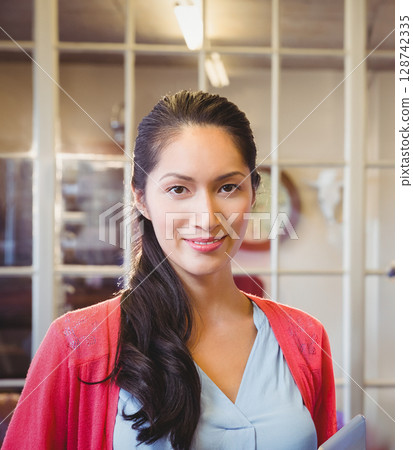 Asian woman standing in office by glass partition holding tablet, smiling with wall clock behind 128742335