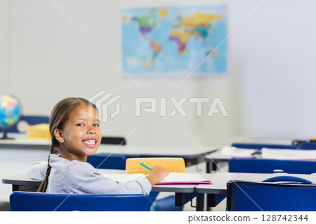 African American elementary school girl writing at desk in classroom with pink notebook, copy space African American elementary school girl writing at desk in classroom with pink notebook, copy space 128742344