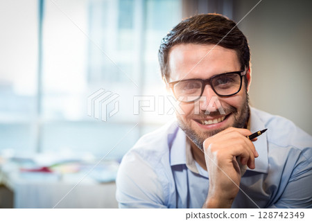 Male worker in early 30s wearing eyeglasses smiling holding black pen at office desk, copy space Male worker in early 30s wearing eyeglasses smiling holding black pen at office desk, copy space 128742349