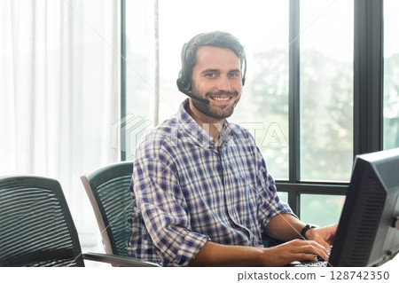 Man typing on keyboard in modern office at floor-to-ceiling windows, smiling with headset Man typing on keyboard in modern office at floor-to-ceiling windows, smiling with headset 128742350