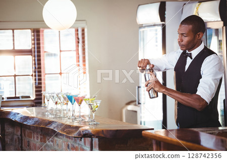 African American bartender shaking cocktail shaker behind wood bar with martini glasses, copy space 128742356