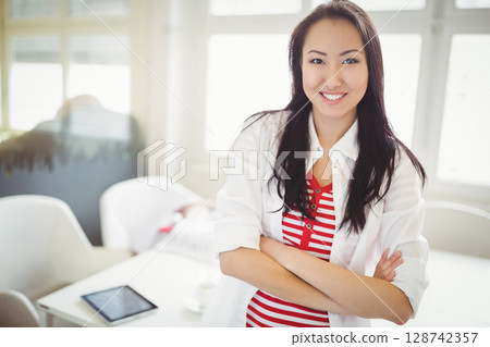 Asian woman folding arms and smiling in bright office with tablet, coffee cup, copy space 128742357