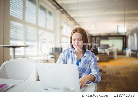 Teenage girl working on silver laptop at open-plan office near windows, with white desk, coffee cup 128742358