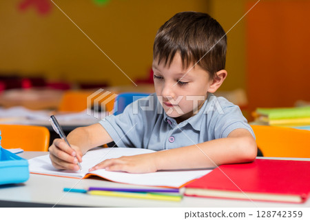 Boy writing at school desk in elementary classroom, with pen open notebook red folder pencil case 128742359